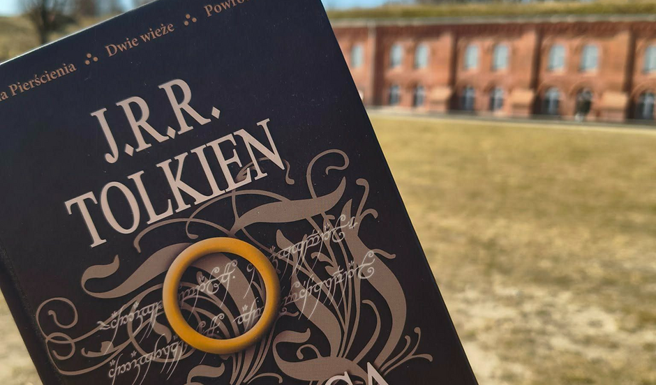 Zdjęcie przedstawia polskie wydanie książki J.R.R. Tolkiena, na której okładce położono żółty pierścień nawiązujący do słynnego Jedynego Pierścienia. W tle widoczny jest zabytkowy, ceglany budynek, co nadaje całości klimatyczny, historyczny charakter. 