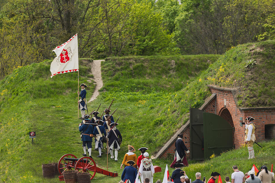 Zdjęcie przedstawia grupę rekonstrukcyjną w historycznych mundurach i strojach z epoki, maszerującą z flagą i armatą na tle trawiastych fortyfikacji. Wydarzenie odbywa się przy ceglanym wejściu do schronu lub kazamaty, przyciągając uwagę zgromadzonych widzów.
