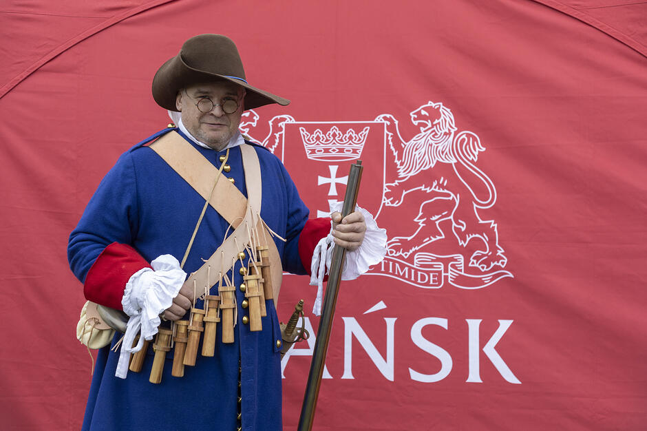 Mężczyzna na zdjęciu to rekonstruktor w stroju muszkietera z XVII wieku, ubranym w charakterystyczny niebieski kaftan z czerwonymi wyłogami oraz pas z dwunastoma drewnianymi pojemnikami na proch, nazywanymi dwunastoma apostołami . Postać pozuje na tle czerwonej flagi z herbem Gdańska, co sugeruje powiązanie z lokalnymi grupami historycznymi, takimi jak Garnizon Gdańs