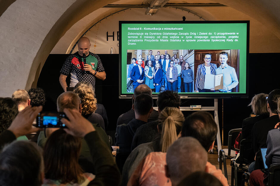 Mężczyzna przemawia na mównicy, obok ekran.