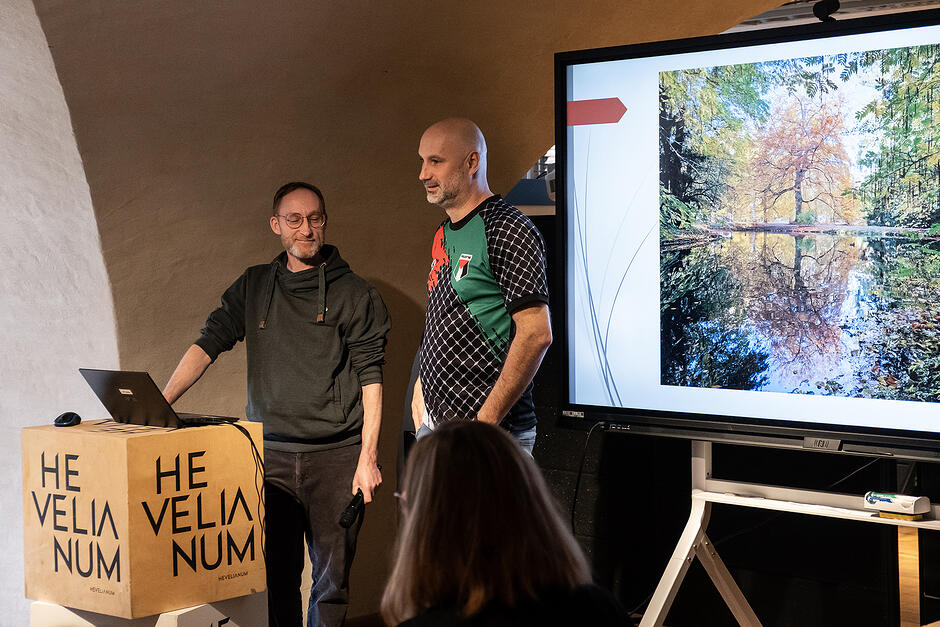 Dwóch mężczyzn stoi na mównicy w sali konferencyjnej, obok stoi ekran.