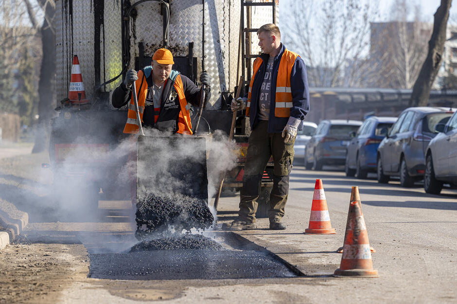 dwóch robotników w pomarańczowych kamizelkach pracuje na ulicy, jeden przewraca taczkę z gorącym asfaltem drugi stoi obok