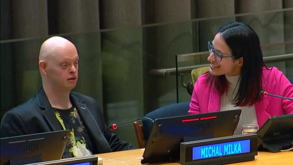 Po lewej stronie siedzi łysy mężczyzna, po prawej kobieta, która uśmiecha się do niego. Oboje siedzą w sali, mówią do mikrofonu