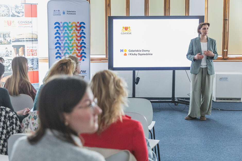 na zdjęciu kobieta w szarej marynarce, stoi i mówi do audytorium