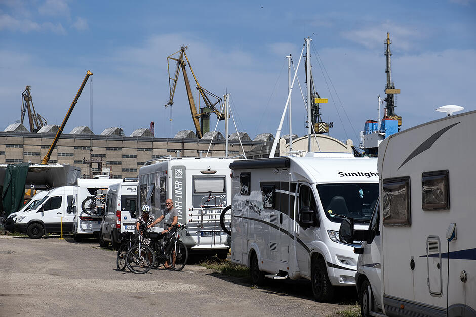 Rząd kamperów zaparkowanych na terenie stoczni, w tle hala stoczniowa i dźwigi. Przy kamperze dwie osoby z rowerami.