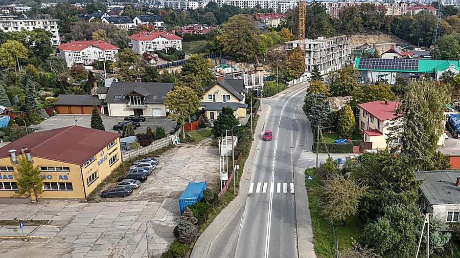 Zdjęcie z lotu ptaka, widok na wąską ulicę w mieście, po której jedzie jeden czerwony samochód. Po bokach ulicy nieduża zabudowa i zieleń miejska.