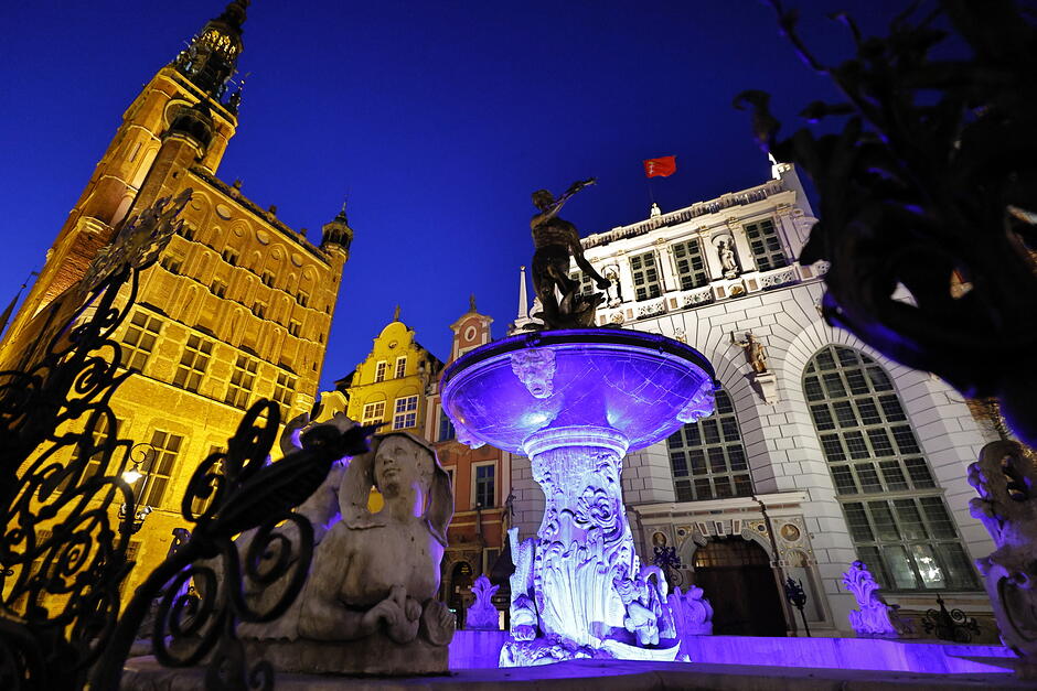 Fontanna w mieście z rzeźbą Neptuna podświetlona na kolor fioletowy. Wokół renesansowa zabudowa.