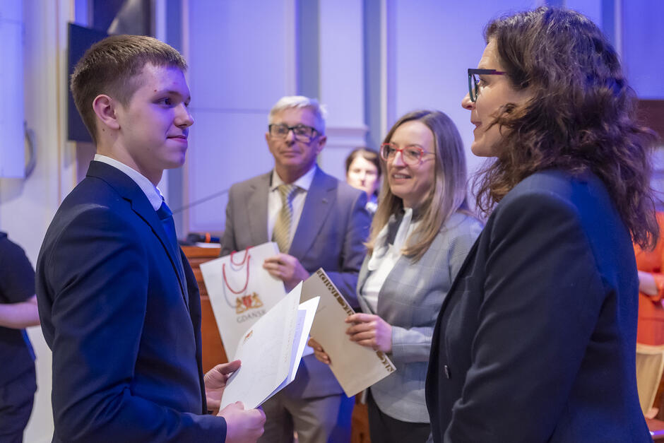 na zdjęciu dwie dorosłe kobiety i mężczyzna, jedna z kobiet gratuluje chłopakowi stojącemu naprzeciwko niej