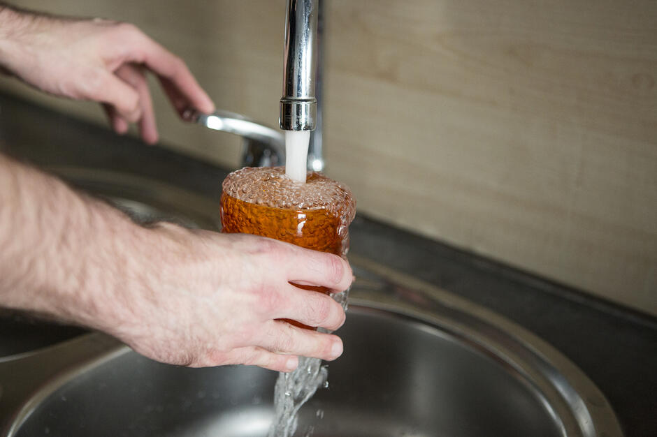 na zdjęciu dwie męskie ręce, owłosione, jedna trzyma pomarańczową szklankę pod kranem z wodą