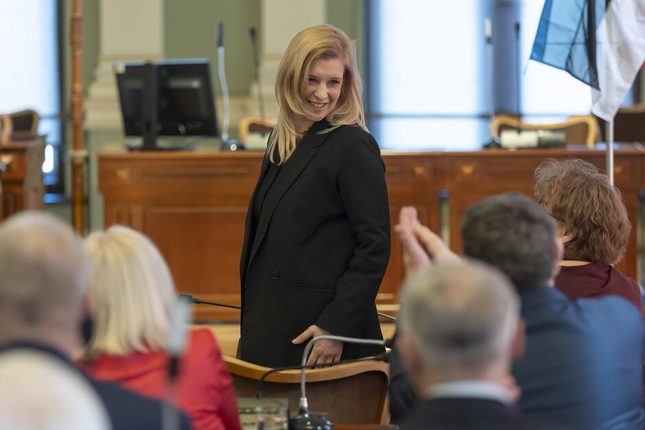 Młoda kobieta stoi w reprezentacyjnej sali i uśmiechając się odwraca się w stronę siedzących w ławach osób.