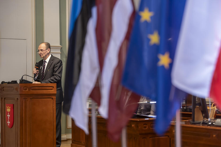 Mężczyzna stoi na mównicy w reprezentacyjnej sali, na pierwszym planie flagi kilku państw.