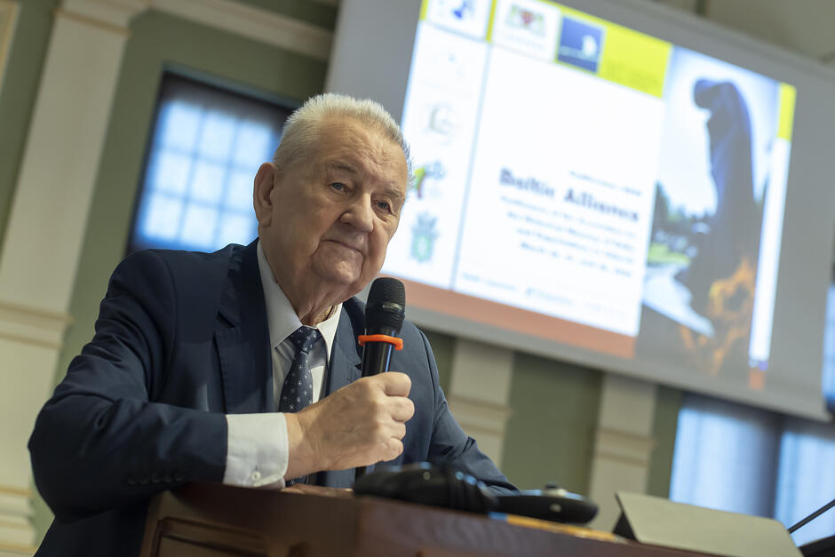 Mężczyzna w starszym wieku przemawia na mównicy w reprezentacyjnej sali.