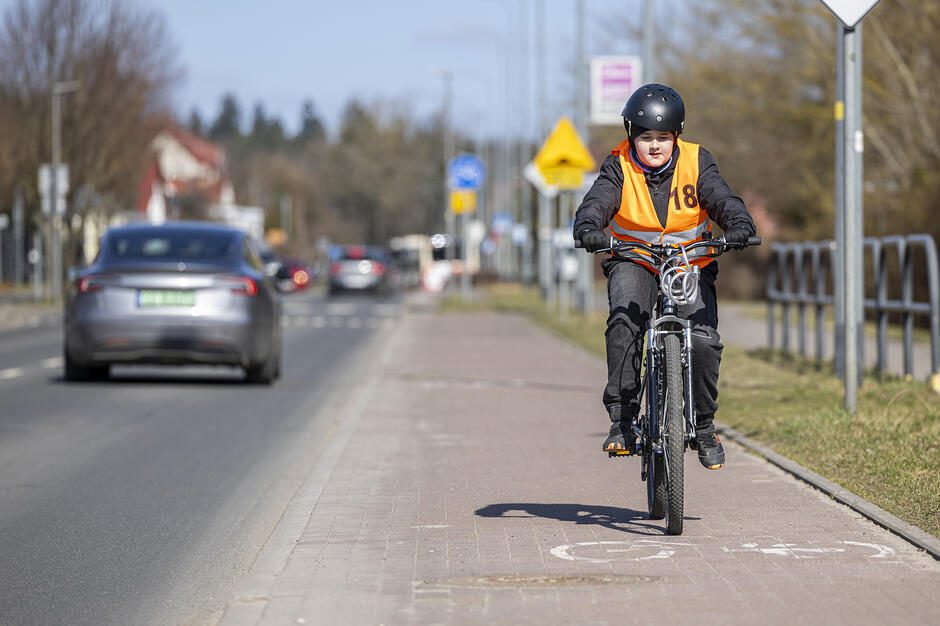 Zdjęcie przedstawia młodego rowerzystę w kasku i odblaskowej kamizelce, który porusza się wyznaczoną ścieżką rowerową wzdłuż jezdni. W tle widać jadące samochody oraz infrastrukturę miejską, co podkreśla znaczenie bezpieczeństwa w ruchu drogowym.