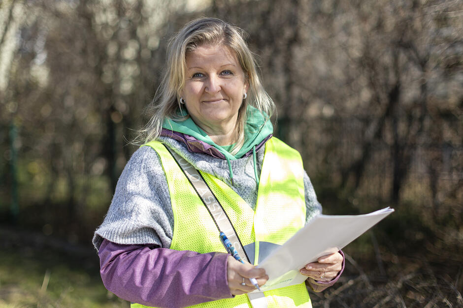 Zdjęcie przedstawia uśmiechniętą kobietę w odblaskowej kamizelce, która trzyma w rękach dokumenty oraz długopis. Postać znajduje się na zewnątrz, na tle rozmytych drzew, co sugeruje pracę w terenie lub prowadzenie działań organizacyjnych. 