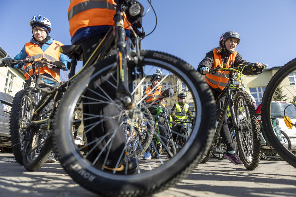 Zdjęcie przedstawia grupę dzieci przygotowujących się do jazdy na rowerach, uchwyconą z niskiej perspektywy przez koło jednego z pojazdów. Wszyscy uczestnicy mają na sobie kaski oraz odblaskowe kamizelki, co wskazuje na zorganizowane zajęcia z bezpieczeństwa w ruchu drogowym.