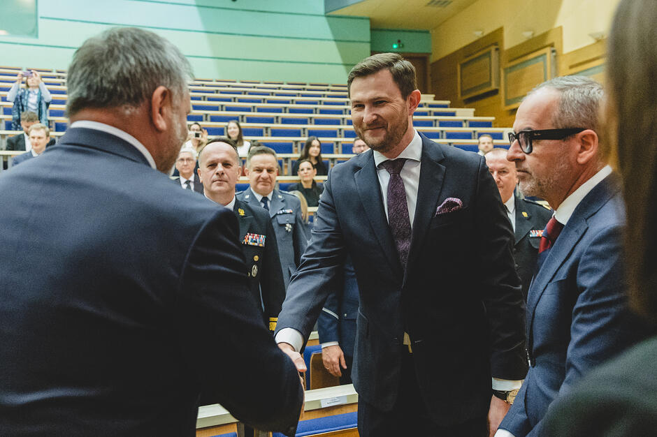 W auli uniwersytetu witają się dwaj mężczyźni przez podanie ręki, obok stoi inny mężczyzna, za nimi ludzie.