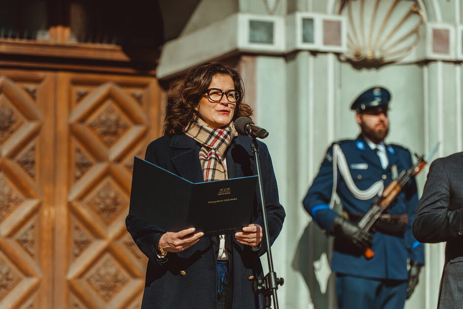 Kobieta przemawia przed wejściem do renesansowego budynku. Za nią stoi na warcie mężczyzna w mundurze i z karabinem.