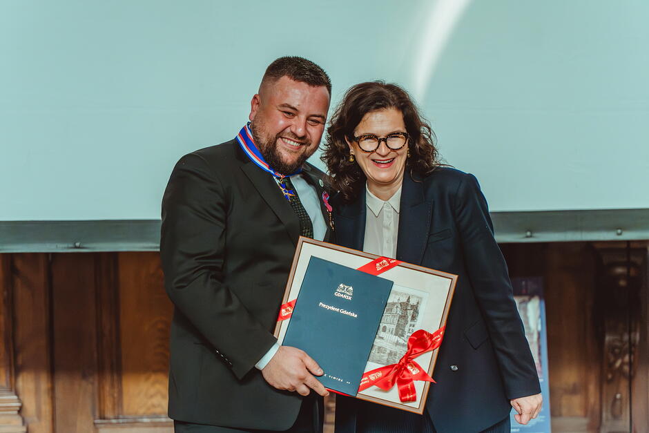 Kobieta i mężczyzna pozują do zdjęcia trzymając oprawioną w ramkę grafikę, uśmiechają się.