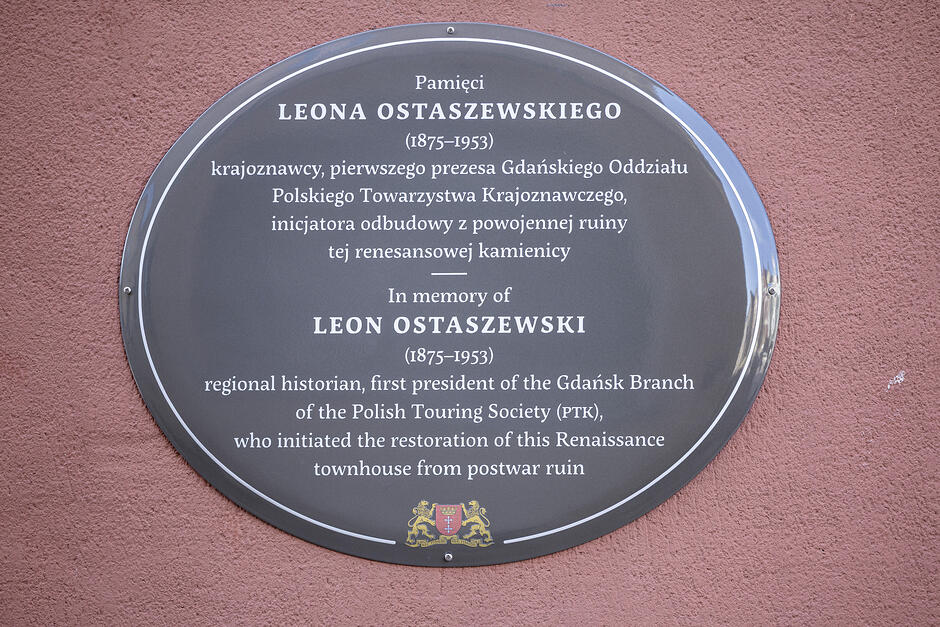 Zdjęcie metalowej emaliowanej tablicy o owalnym kształcie. Jest przymocowana do ściany budynku