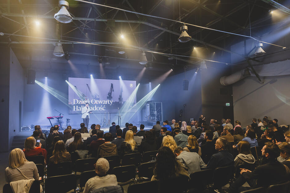 na zdjęciu przyciemniona sala z reflektorami i siedzącymi ludźmi, widać oświetloną scenę na której stoi prowadzący