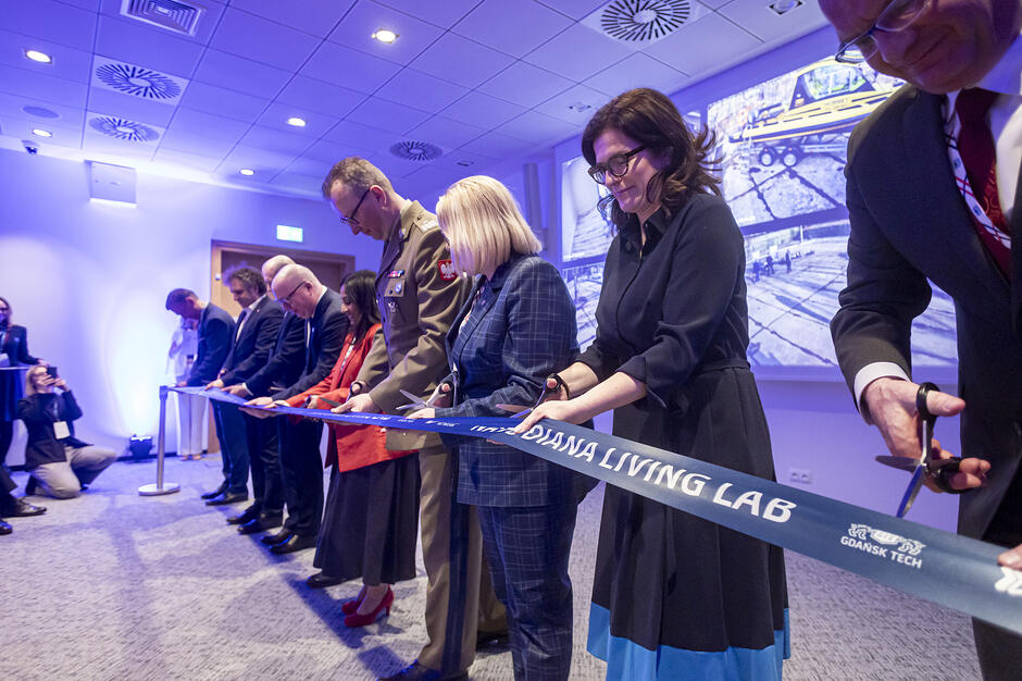 Na zdjęciu widać grupę osób uczestniczących w uroczystym otwarciu „DIANA Living Lab” na Politechnice Gdańskiej. Zgromadzeni, wśród których są przedstawiciele uczelni oraz wojska, wspólnie przecinają niebieską wstęgę z logotypem placówki. 