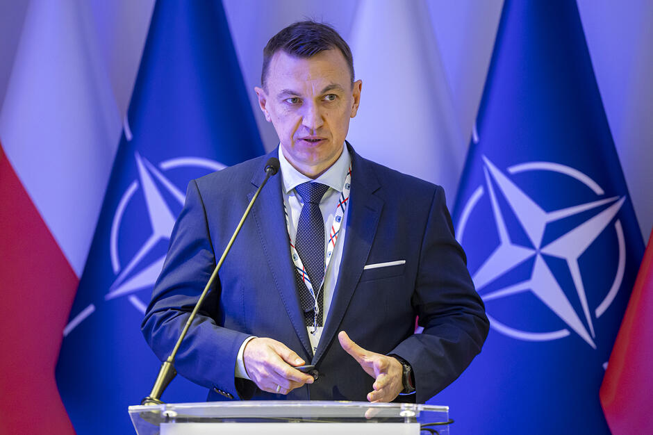 Na zdjęciu widać mężczyznę przemawiającego podczas oficjalnego wydarzenia. Stoi on przy pulpicie na tle flag Polski oraz NATO, trzymając w dłoni pilot do prezentacji. 