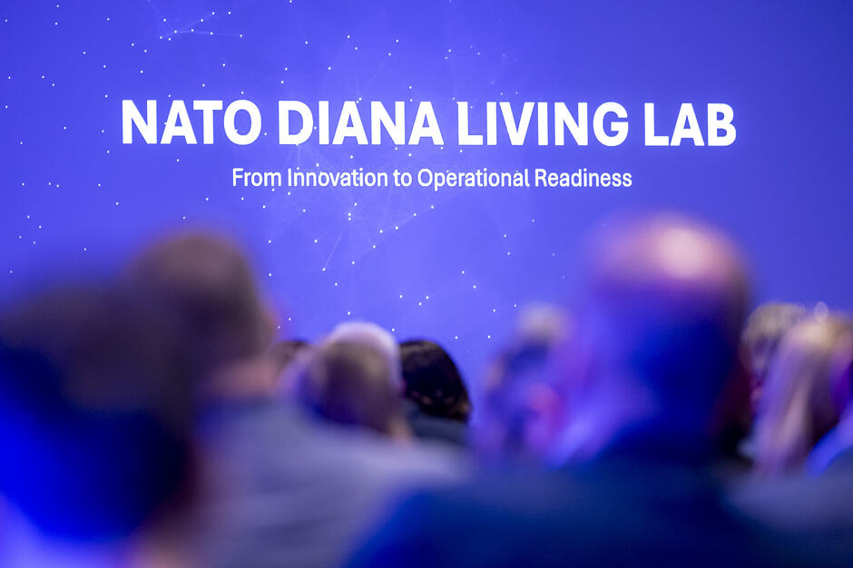 Na grafice widoczny jest duży napis „NATO DIANA LIVING LAB” wraz z hasłem o przechodzeniu od innowacji do gotowości operacyjnej. Napis znajduje się na niebieskim tle z motywem siatki połączeń, a na pierwszym planie widać rozmyte sylwetki osób uczestniczących w wydarzeniu.