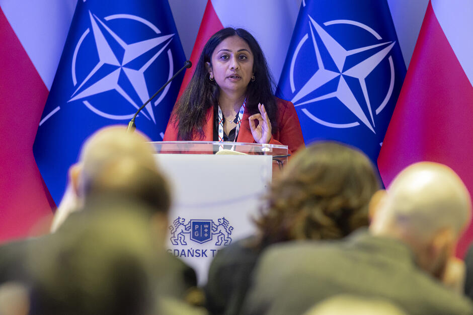 Kobieta przemawia zza mównicy ozdobionej herbem Gdańska. W tle znajdują się flagi Polski oraz NATO, a na pierwszym planie widoczne są sylwetki słuchaczy uczestniczących w wydarzeniu.