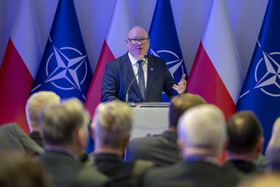 Na zdjęciu widać mężczyznę w okularach przemawiającego zza mównicy podczas oficjalnego wydarzenia, na co wskazują flagi Polski i NATO w tle. Przed nim siedzi liczna grupa słuchaczy, których sylwetki są widoczne na pierwszym planie w lekkim rozmyciu.