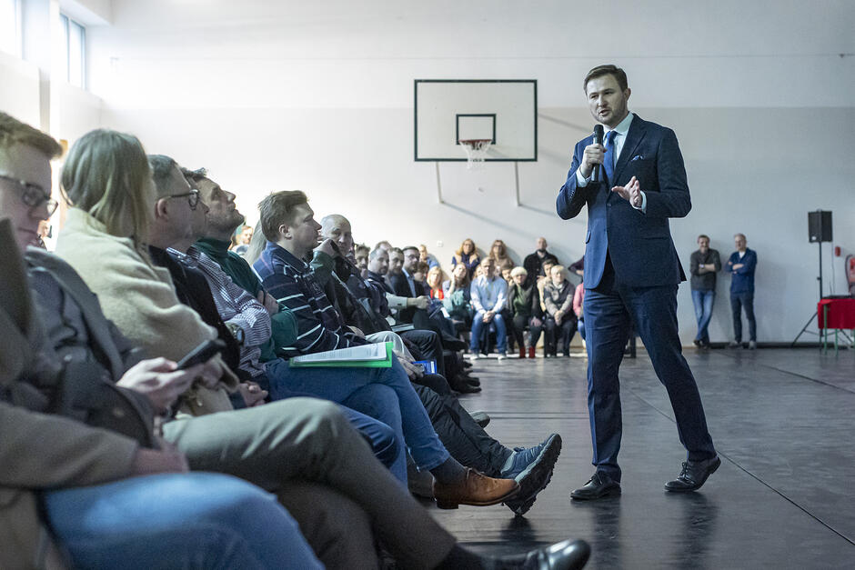 Mężczyzna z mikrofonem mówi i chodzi przed osobami siedzącymi na krzesłach, w rzędach
