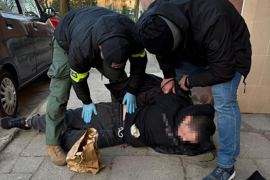 Dwóch mężczyzn w czarnych kurtkach i opaskach na ramieniu z napisem Policja schyla się do leżącego na ziemi mężczyzny i trzyma go, obok leży papierowa torba z pieniędzmi. Obok stoi samochód.