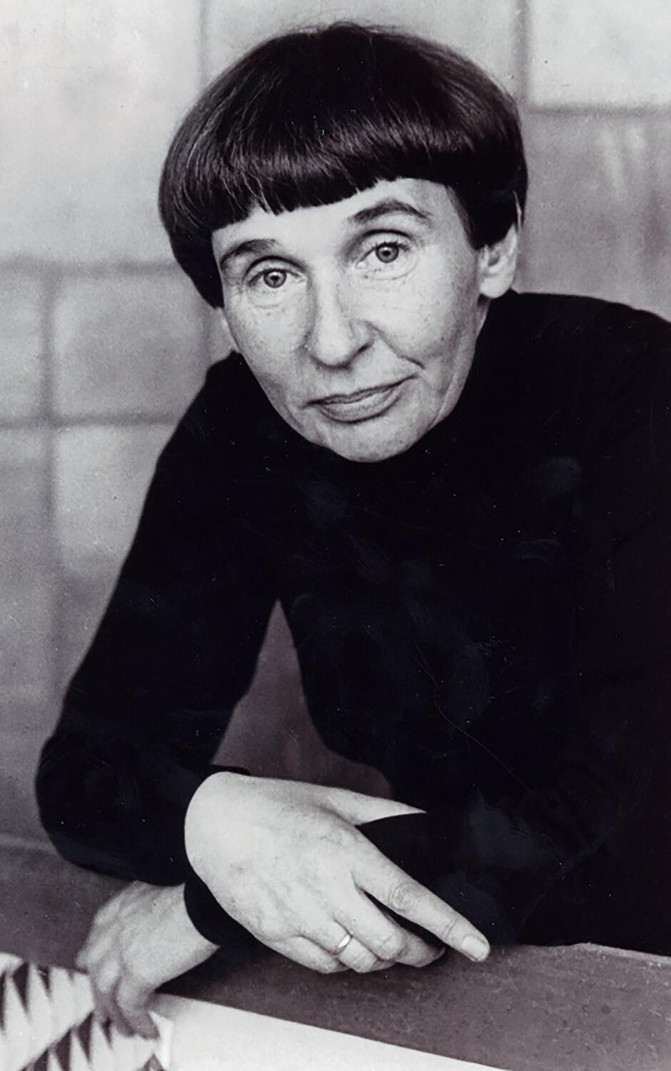 Czarnobiały portret kobiety w średnim wieku z czarnymi, krótkimi włosami z grzywką. Kobieta patrzy w obiektyw, delikatnie uśmiecha i podpiera się rękoma o blat biurka.