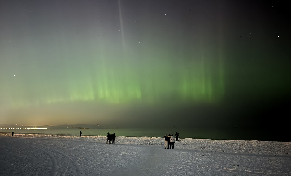Zdjęcie przedstawia grupę ludzi obserwujących zjawiskową zorzę polarną, która rozświetla nocne niebo intensywnymi, zielonymi smugami. Scena rozgrywa się na ośnieżonym wybrzeżu, gdzie światła zjawiska odbijają się w tafli wody, tworząc magiczny i mroźny krajobraz. 