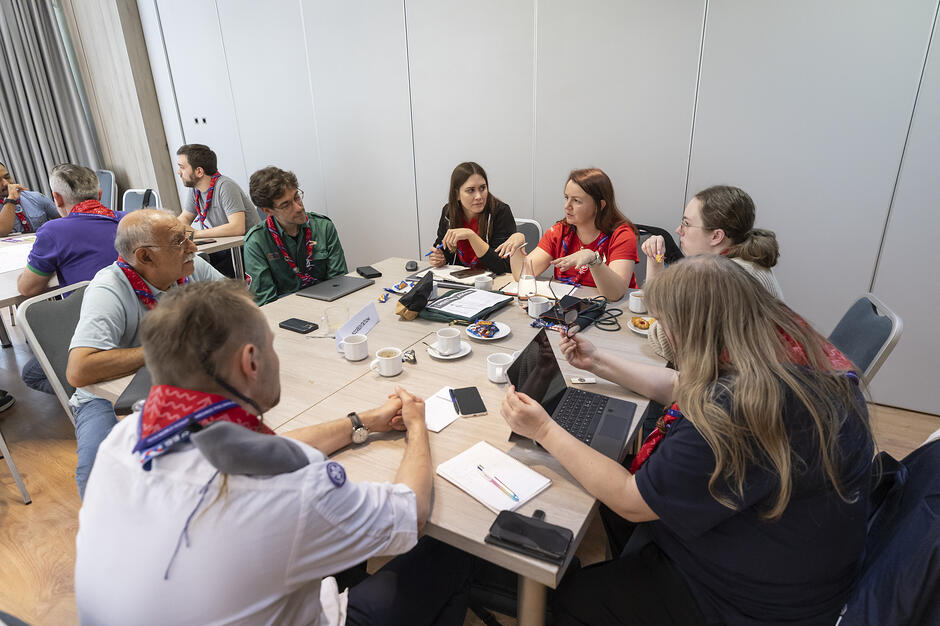 Zdjęcie przedstawia grupę osób siedzących przy stole w jasnym pomieszczeniu, prowadzących rozmowę w atmosferze warsztatów lub spotkania roboczego. Wiele z tych osób ma na sobie harcerskie chusty, a na blacie leżą laptopy, notatniki oraz filiżanki z napojami. 