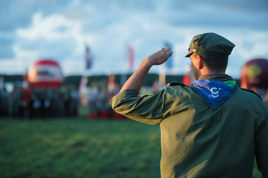 Zdjęcie przedstawia harcerza w mundurze i rogatywce, który salutuje w stronę zgromadzonej w oddali grupy osób. Cała scena rozgrywa się na otwartej przestrzeni podczas słonecznego dnia, a w tle widać powiewające flagi.