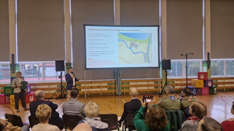 Zdjęcie przedstawia spotkanie informacyjne w sali gimnastycznej, podczas którego na dużym ekranie prezentowany jest harmonogram modernizacji infrastruktury drogowej. Przed ekranem stoi prelegent z mikrofonem, a na widowni zasiadają uczestnicy, wśród których widać osoby w harcerskich mundurach.