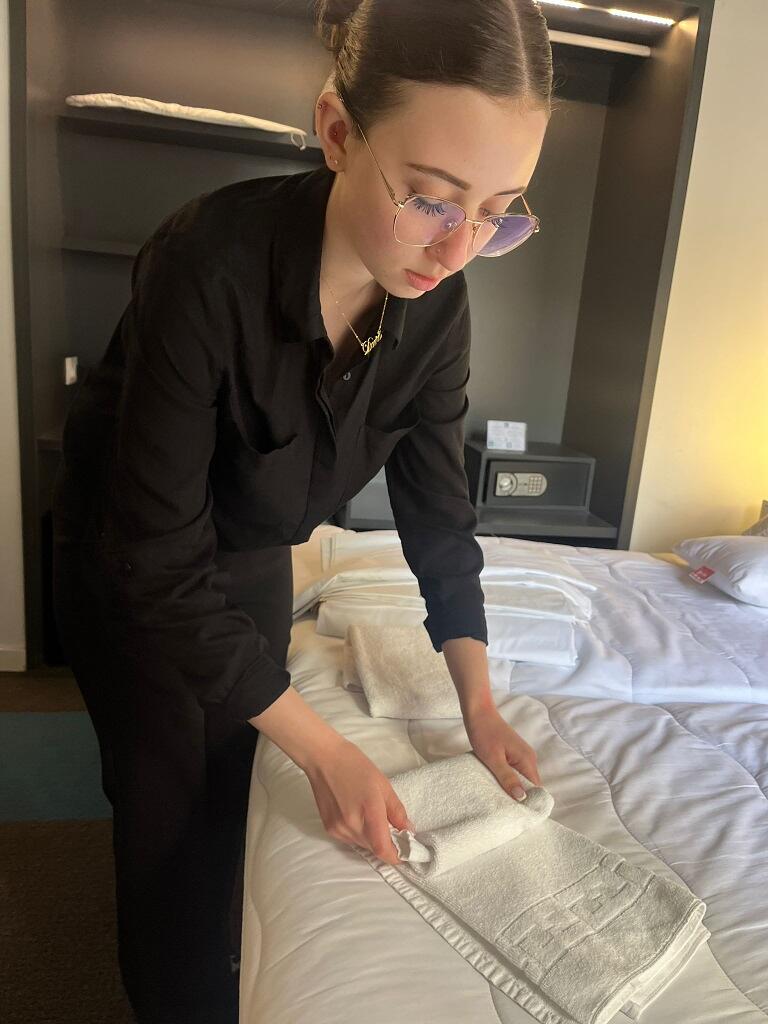 Zdjęcie przedstawia młodą kobietę w czarnym stroju, która z dużym skupieniem ściele łóżko i starannie składa białe ręczniki. Całość odbywa się w nowocześnie urządzonym pokoju hotelowym, wyposażonym w sejf oraz minimalistyczne meble.