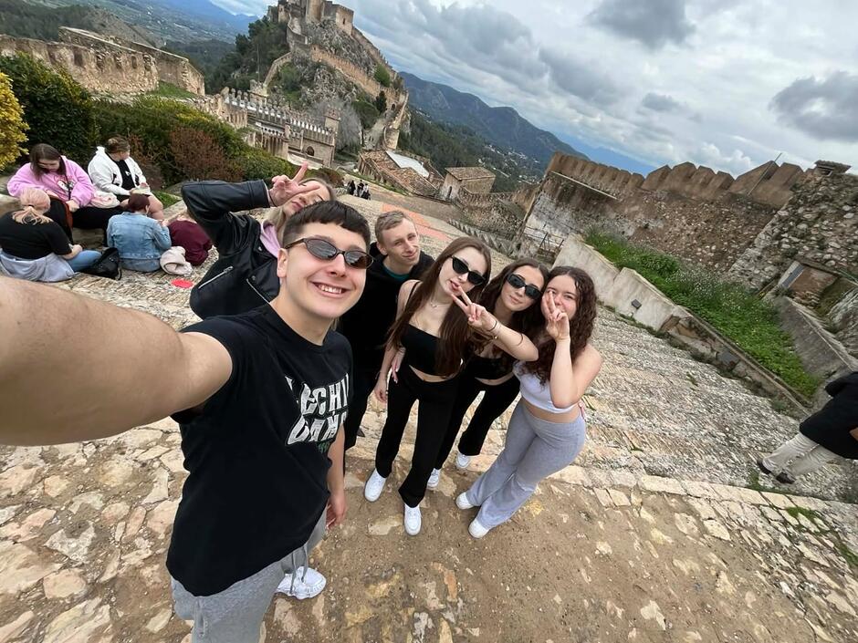 Grupa roześmianych młodych ludzi robi wspólne selfie na tle malowniczych ruin średniowiecznego zamku w Hiszpanii. W tle widać kamienne mury obronne, wieże oraz rozległy górski krajobraz pod lekko zachmurzonym niebem.
