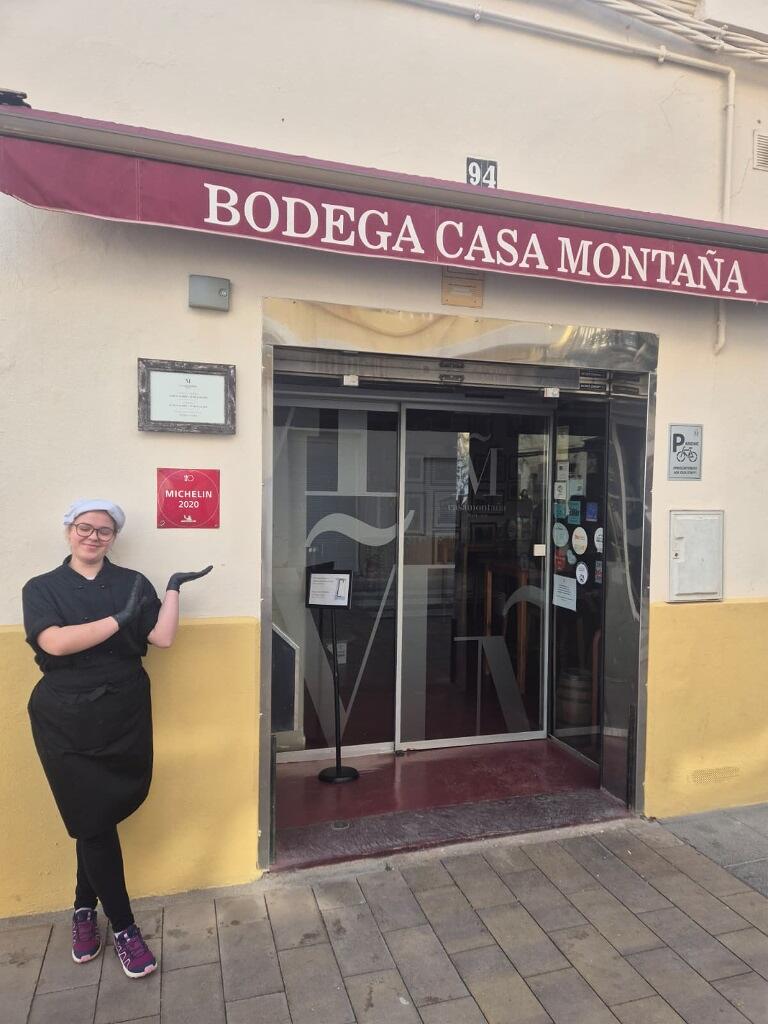 młoda kobieta stoi przy wejściu do restauracji o hiszpańskojęzycznej nazwie i wskazuje na nią