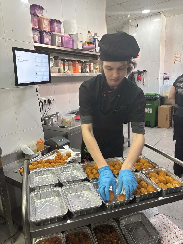 Młody kucharz w czarnym stroju i niebieskich rękawiczkach z uwagą układa smażone przekąski, prawdopodobnie krokiety, w aluminiowych pojemnikach na wynos. Całość dzieje się w profesjonalnej kuchni, gdzie na regałach w tle widać liczne przyprawy, pojemniki oraz ekran systemu zamówień. 