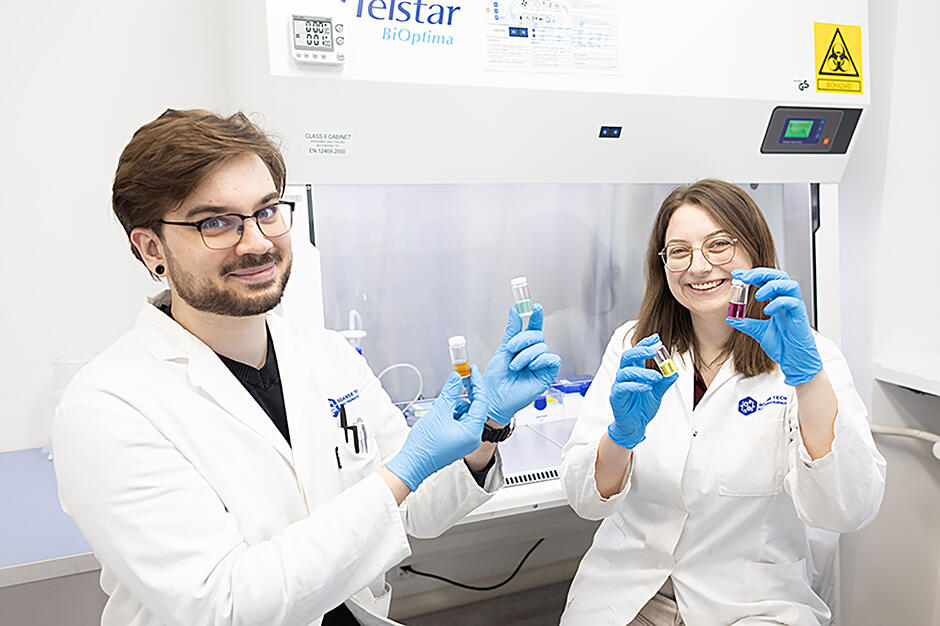 Kobieta i mężczyzna - naukowcy, siedzą w laboratorium w białych fartuchach i pozują do zdjęcia, uśmiechając się i pokazując próbówki. 