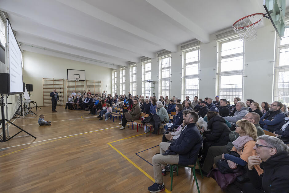 szkolna sala gimnastyczna wypełniona ludźmi, ponad sto osób siedzi na krzesłach, patrzy przed siebie