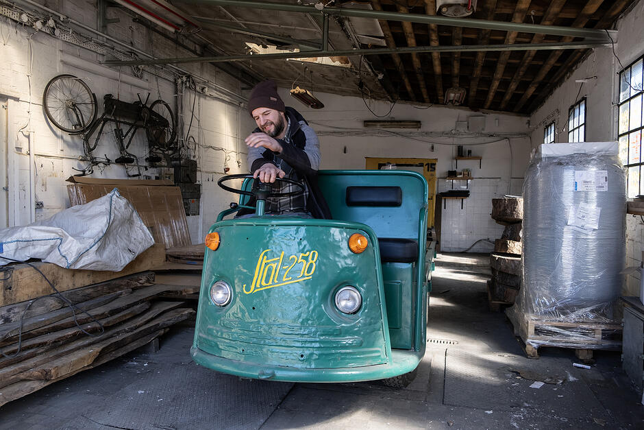 Mężczyzna w czapce siedzi za kierownica wózka akumulatorowego w warsztacie.