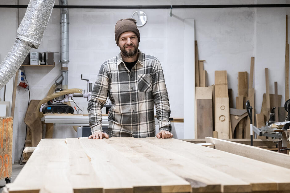 Mężczyzna w czapce i koszuli flanelowej w kratę stoi w stolarni i opiera się rękoma o drewniany blat. Pozując do zdjęcia delikatnie uśmiecha się. 