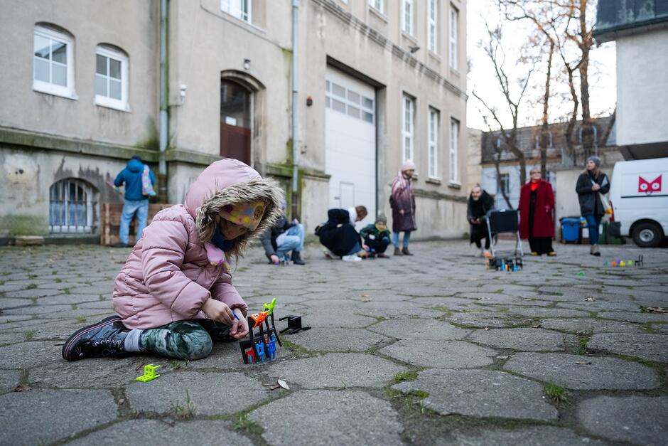 Na zdjęciu widać dziecko w różowej kurtce, które bawi się kolorowymi zabawkami na wybrukowanym, miejskim podwórku. W tle znajduje się grupa dorosłych i dzieci stojących przed starym, wielopiętrowym budynkiem o jasnej elewacji.