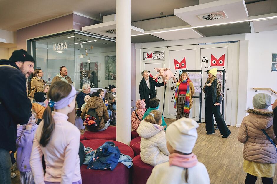 Zdjęcie przedstawia grupę dzieci i dorosłych zgromadzonych w jasnym wnętrzu teatru, gdzie odbywa się pokaz z użyciem pacynki świnki. Widzowie siedzą na czerwonych pufach i obserwują występy artystów na tle biało-różowych dekoracji z motywami kotów.