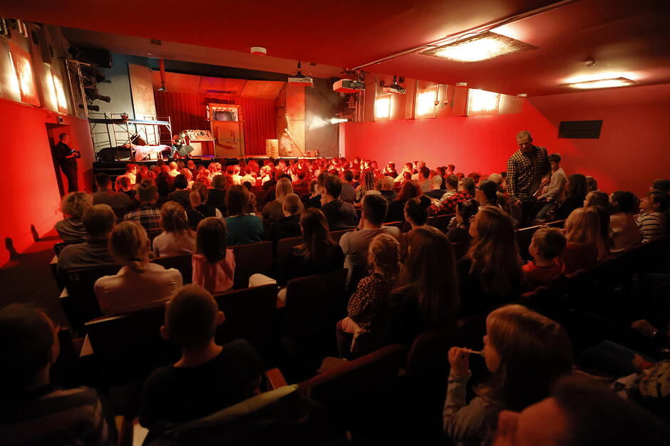 Zdjęcie przedstawia wypełnioną po brzegi widownię teatru, na której dorośli i dzieci wspólnie oglądają spektakl. Cała sala jest spowita nastrojowym, czerwonym światłem, a uwaga wszystkich widzów skupiona jest na oświetlonej scenie.
