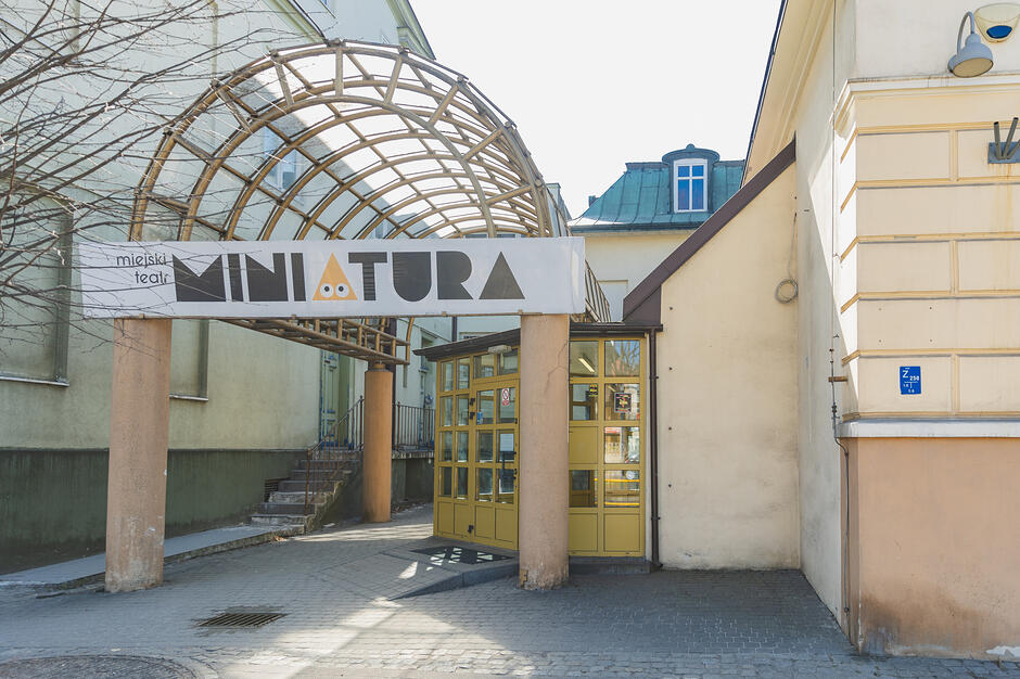 Zdjęcie przedstawia wejście do Miejskiego Teatru Miniatura, nad którym znajduje się biały szyld z nazwą instytucji i charakterystyczną grafiką oczu. Budynek ma jasną elewację, żółte przeszklone drzwi oraz charakterystyczne łukowe zadaszenie z metalowych prętów.

