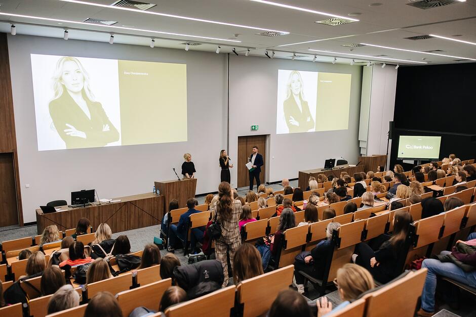 Aula WSB Merito wypełniona uczestnikami wydarzenia - spotkania z Ewą Chodakowską, która stoi przed tłumem i wita się z gośćmi.