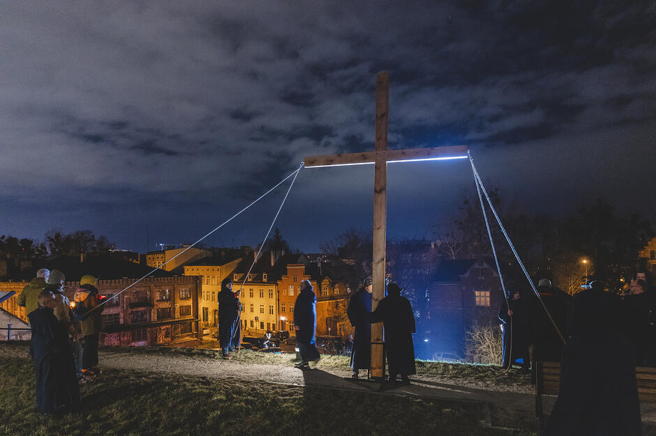 Podnoszenie do pionu dużego krzyża, wokół jest ciemno - krzyż podświetlony jest reflektorem, wokół widać sylwetki osób uczestniczących w procesji